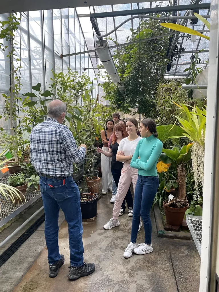 A male presenter talks to students in the greenhouse, surrounded by greenery